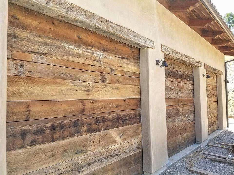 Three garage bays clad in reclaimed timber of varied tone and grain, set
                  into smooth tan stucco walls under a deep timber eave, with black
                  gooseneck barn lights mounted above each opening.