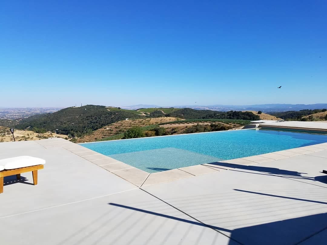 An infinity-edge pool set into a concrete deck on top of a ridge. The
                  water runs to a vanishing edge with nothing beyond it but oak-dotted
                  hills, vineyard rows and open valley to the horizon under a clear sky.
                  A teak lounger sits at the left.