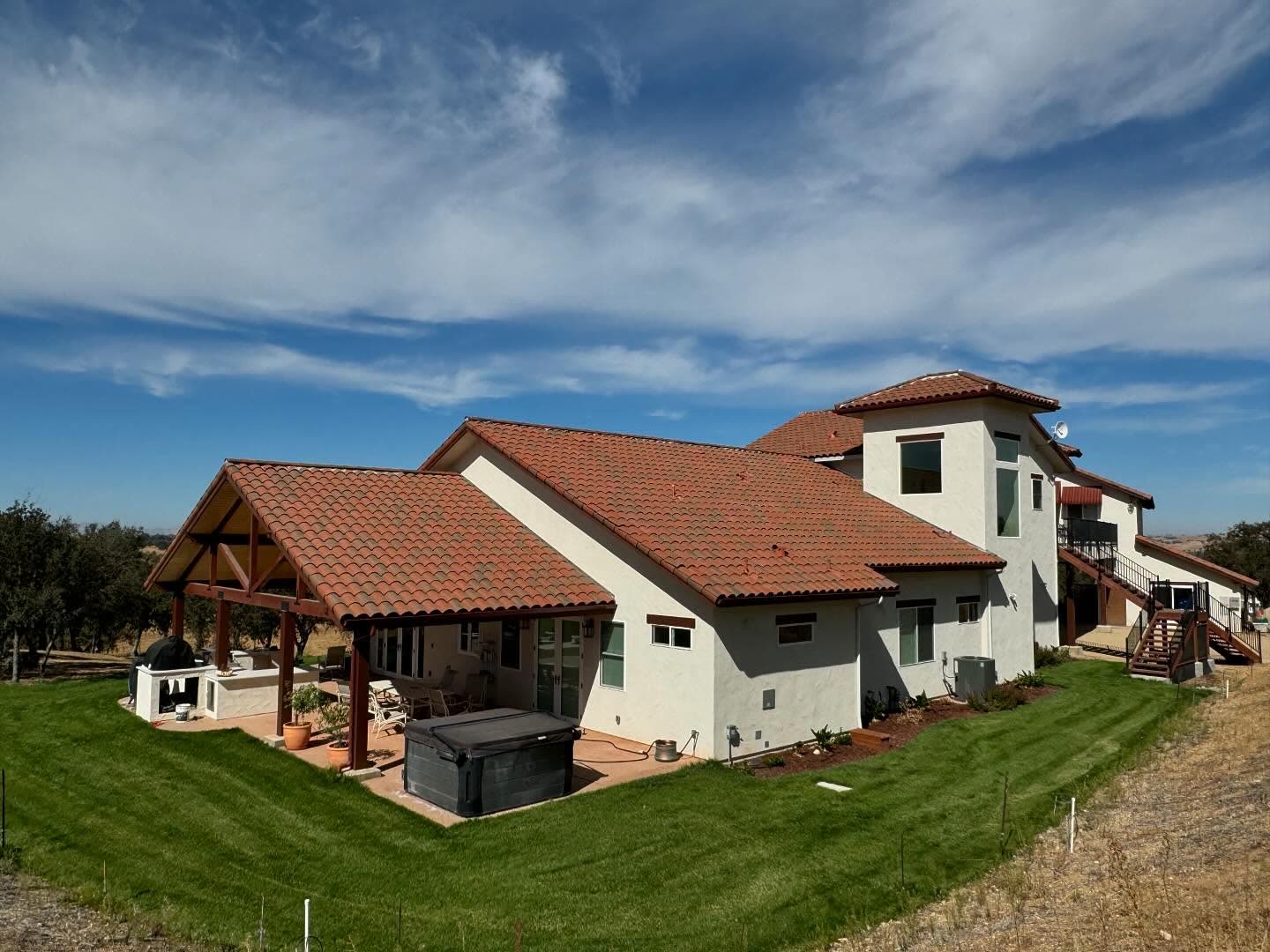 A large single-storey ranch house finished in cream stucco under a terracotta
            barrel tile roof, photographed from the lawn. A timber-framed loggia with
            heavy posts and beams shelters an outdoor kitchen and a sunken spa, a second
            wing with a balcony runs up the slope behind, and oaks and open hills sit
            beyond.