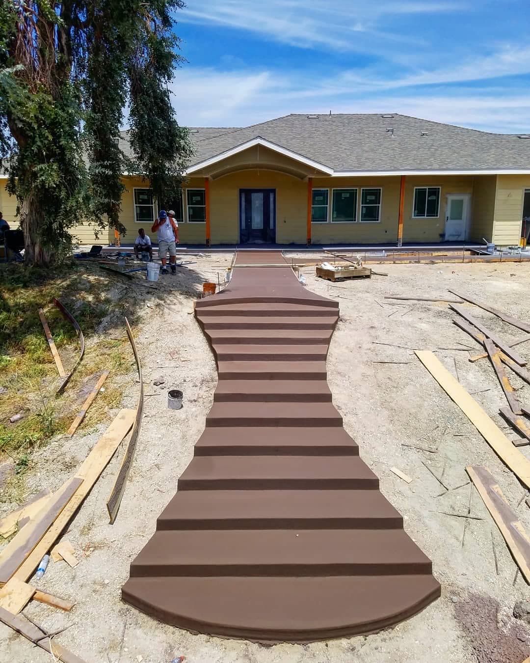 A wide curved concrete walkway, freshly finished in a warm brown, sweeping
                  from the foreground up to the covered entry of a yellow craftsman-style
                  house. Forms, lumber and buckets lie either side and two crew members
                  work near the porch under a bright sky.